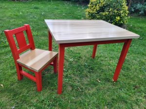 Child-sized wooden table and chair set with natural wood tops and vibrant red legs, placed on a grassy lawn with a lush garden backdrop—perfect for outdoor play or creative activities.