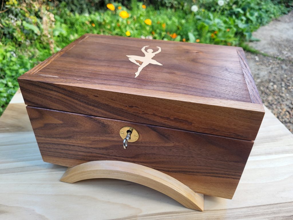 A wooden box with a carved symbol on the top, placed on a light wooden surface, surrounded by greenery and flowers.