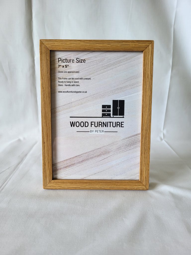 A rectangular wooden picture frame displayed on a white fabric background. The frame encloses a label that reads ‘WOOD FURNITURE BY PETER’ with a logo. There is additional text providing details about the product’s size and material, which are not fully legible in the image. The natural wood grain is visible, giving the frame a rustic appearance.