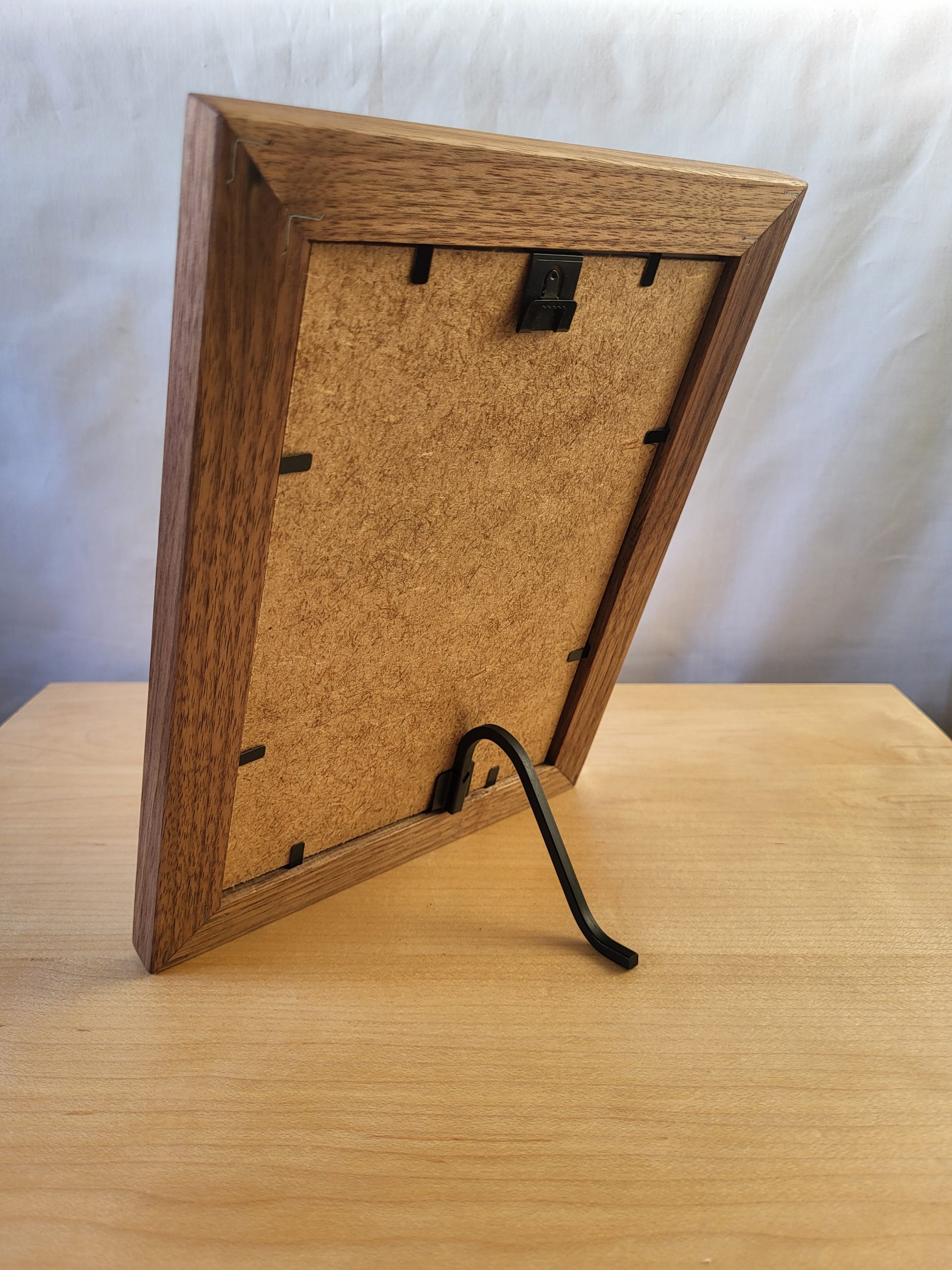 Back view of a handcrafted wooden picture frame resting on a natural wood surface, supported by a sleek black metal stand. The fiberboard backing is secured with black swivel tabs, showcasing thoughtful design and durable craftsmanship.