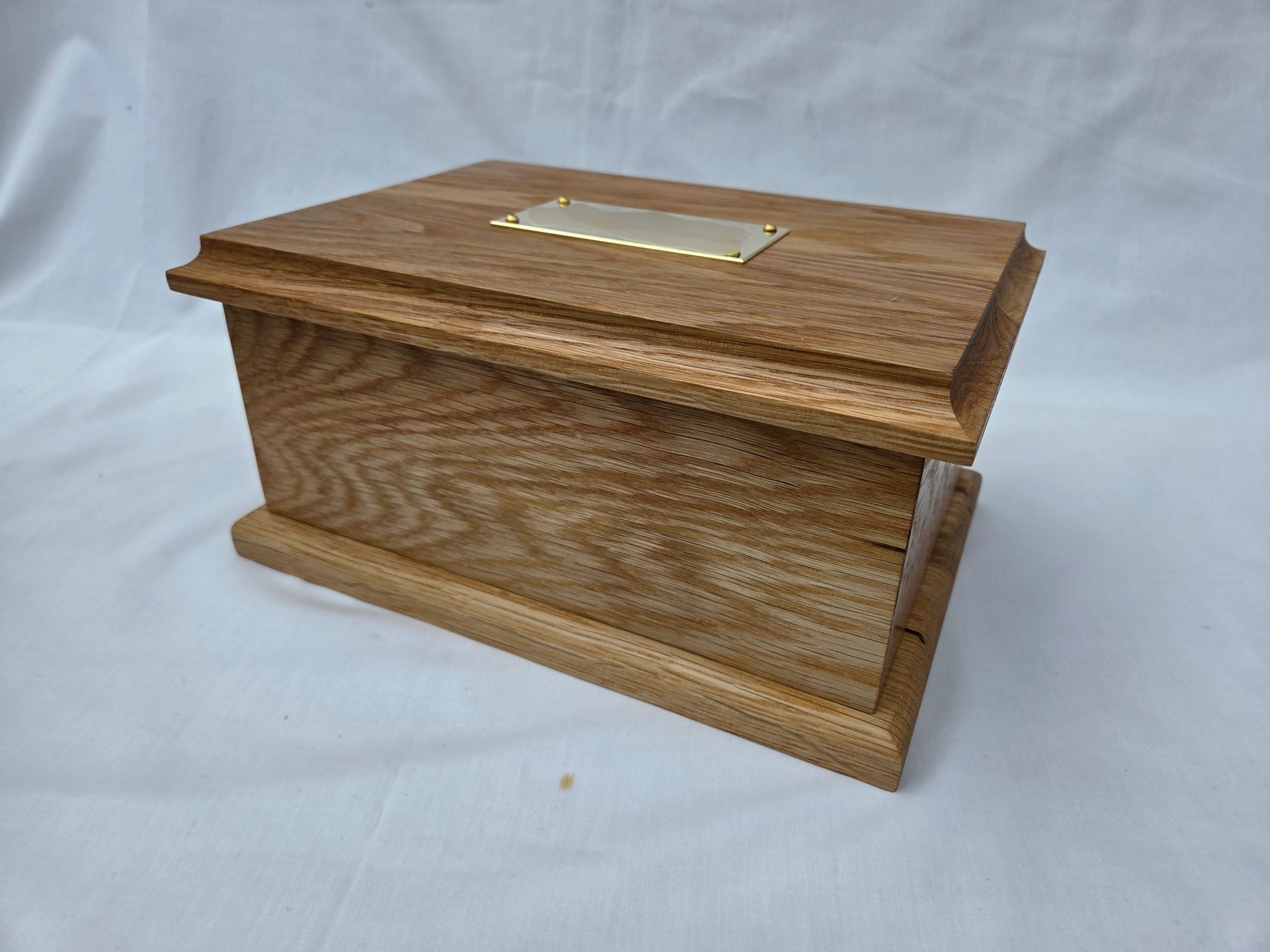 Handcrafted wooden ashes casket box with a polished oak finish, beveled edges, and a raised lid featuring a blank brass nameplate. Displayed on white fabric for presentation, ideal for use as a dignified urn.