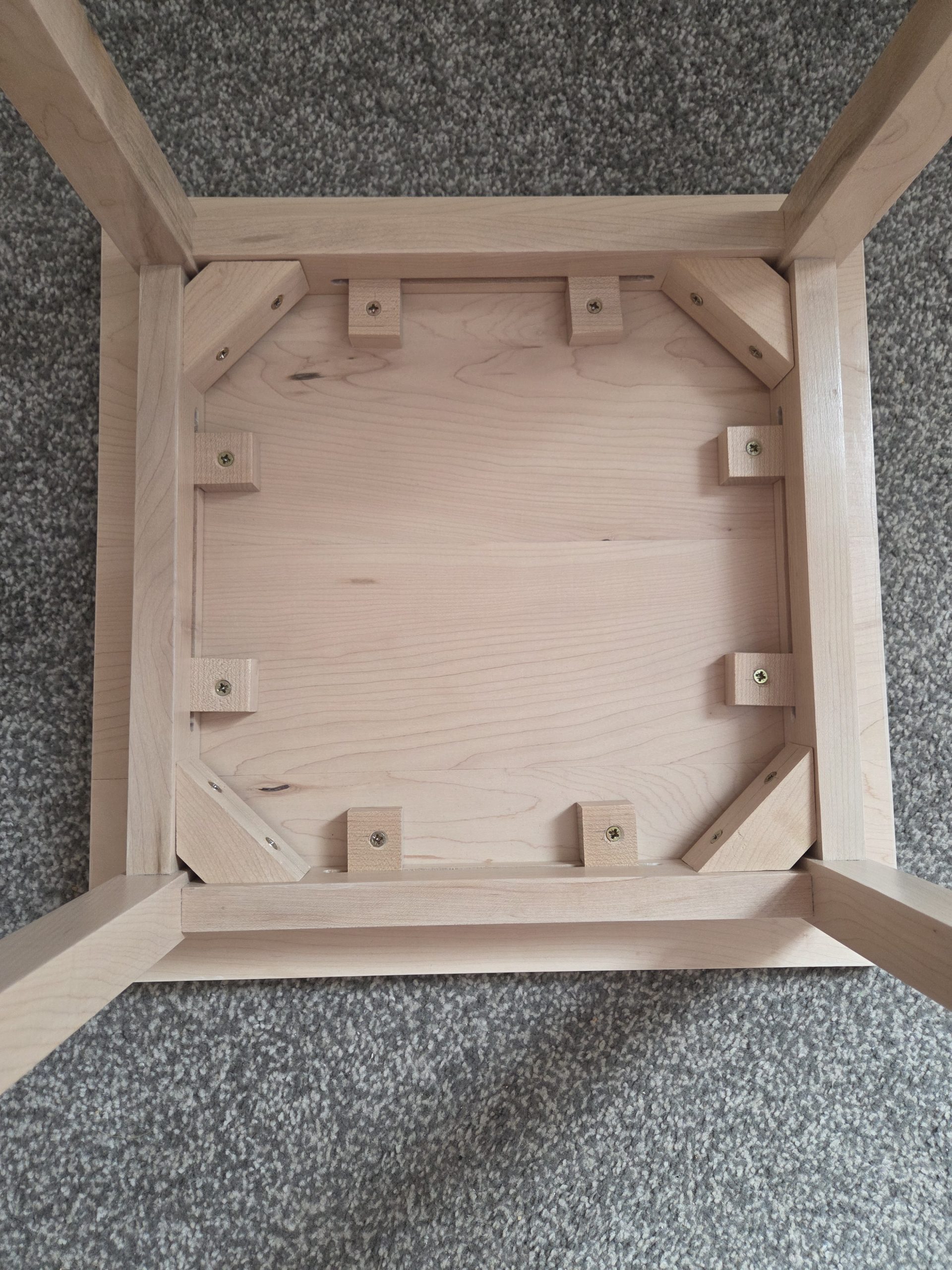 Underside of a handcrafted wooden table, showcasing solid joinery and artisan construction. Four legs are reinforced with triangular corner braces and rectangular support blocks, all precisely fastened with screws for lasting stability. A testament to durable, thoughtful design and expert woodworking.