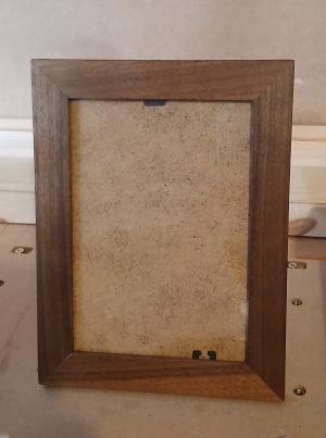A handcrafted walnut picture frame with smooth, dark brown finish and visible wood grain, standing upright on a workshop surface. The frame holds a plain MDF backing board secured by small black clips at the top and bottom