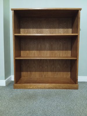 Handmade solid oak bookcase with three vertical compartments, positioned against a light wall and carpeted floor. The bookcase features visible wood grain, smooth satin finish, and sturdy construction with thick side panels and a solid base.