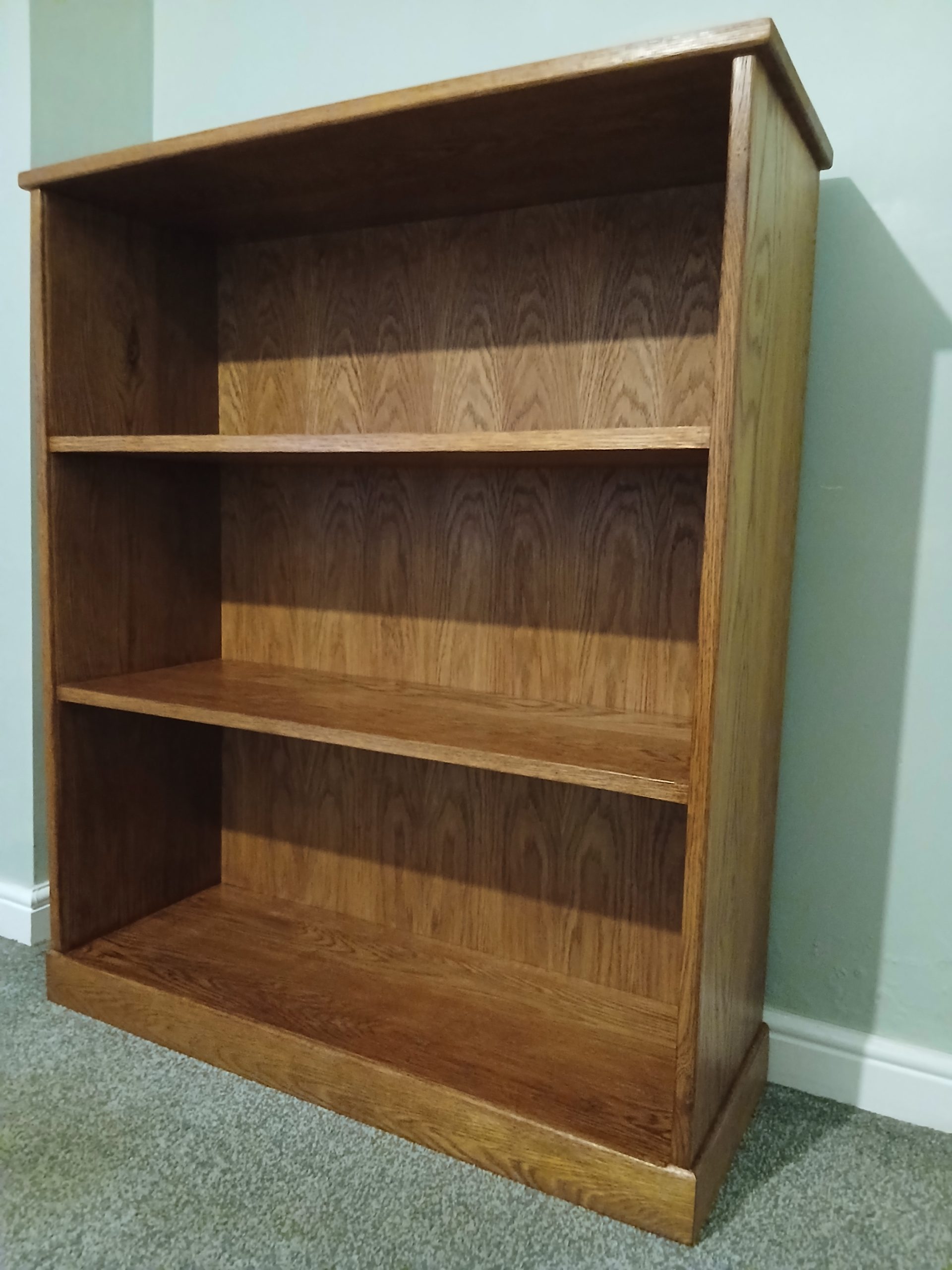 Handcrafted solid oak bookcase with three vertical compartments, positioned against a light wall and carpeted floor. The bookcase features visible oak grain, smooth satin finish, and sturdy joinery with thick side panels and a solid top and base.