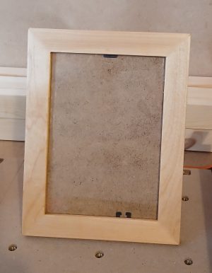 A light natural wood picture frame with a smooth, simple design stands upright on a workbench. The frame holds a plain MDF backing board visible through a glass front, secured by small black clips at the top and bottom. The background shows a neutral workshop setting with wooden boards and a beige wall.