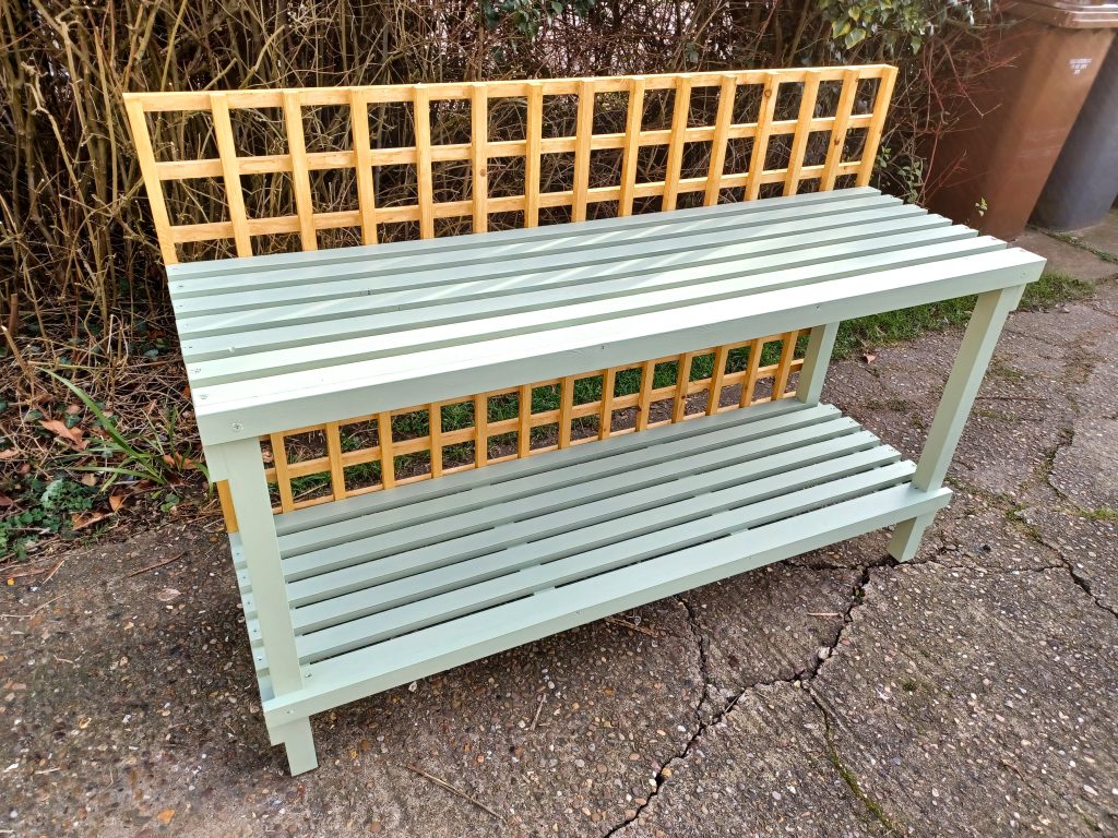 A light sage green wooden garden potting bench with a slatted worktop and a matching lower shelf. The bench features a natural unpainted wood lattice trellis attached along the backrest and between the two levels. It sits outdoors on a paved concrete ground in front of garden foliage.
