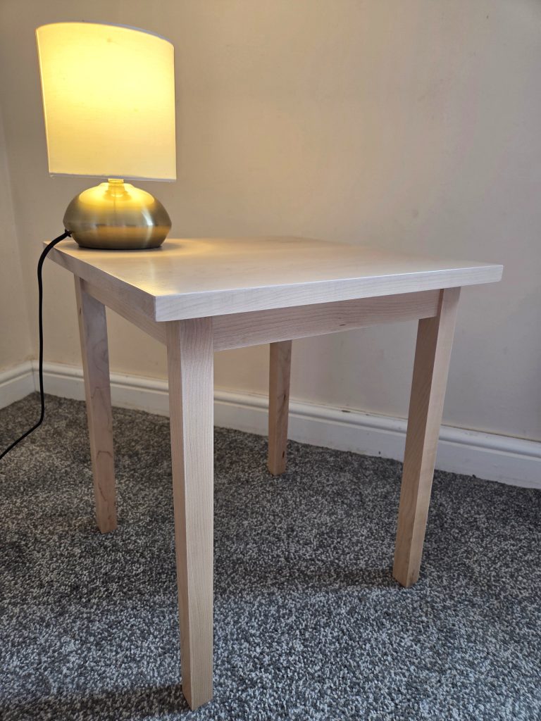 A light-colored solid maple side table sitting on a textured grey carpet in the corner of a room. On top of the table rests a small, illuminated desk lamp featuring a rounded, metallic brass base and a warm white cylindrical shade.