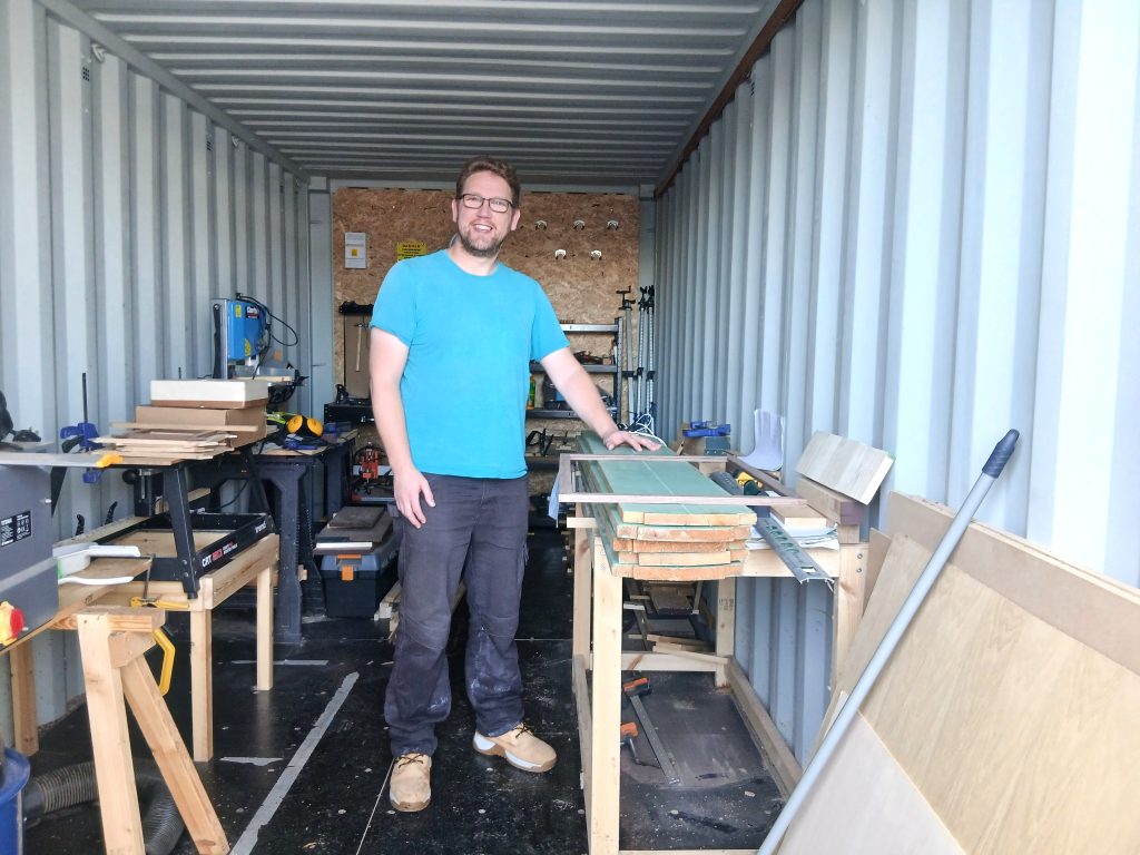 A man wearing glasses, a bright blue t-shirt, and dark trousers standing inside a workshop converted from a shipping container. He is smiling and resting one hand on a wooden workbench stacked with timber and a picture frame. The container's corrugated walls surround the space, which is equipped with woodworking tools, machinery, and wood supplies arranged along the sides.