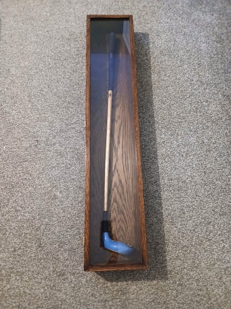 Top-down view of a slim, vertical rectangular display case crafted from dark stained oak wood with visible grain. The case features a clear glass front panel showcasing a golf club with a light wooden shaft, black grip, and a blue clubhead, resting on a textured grey carpet.