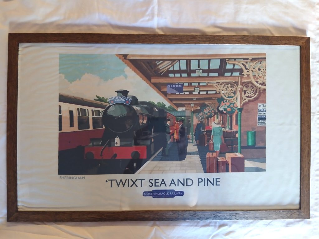 Front-facing view of a wide rectangular dark oak wood frame lying on a white textured background. The frame encloses a vintage North Norfolk Railway art print depicting a steam train at Sheringham station platform with passengers and luggage, featuring the title "'TWIXT SEA AND PINE".