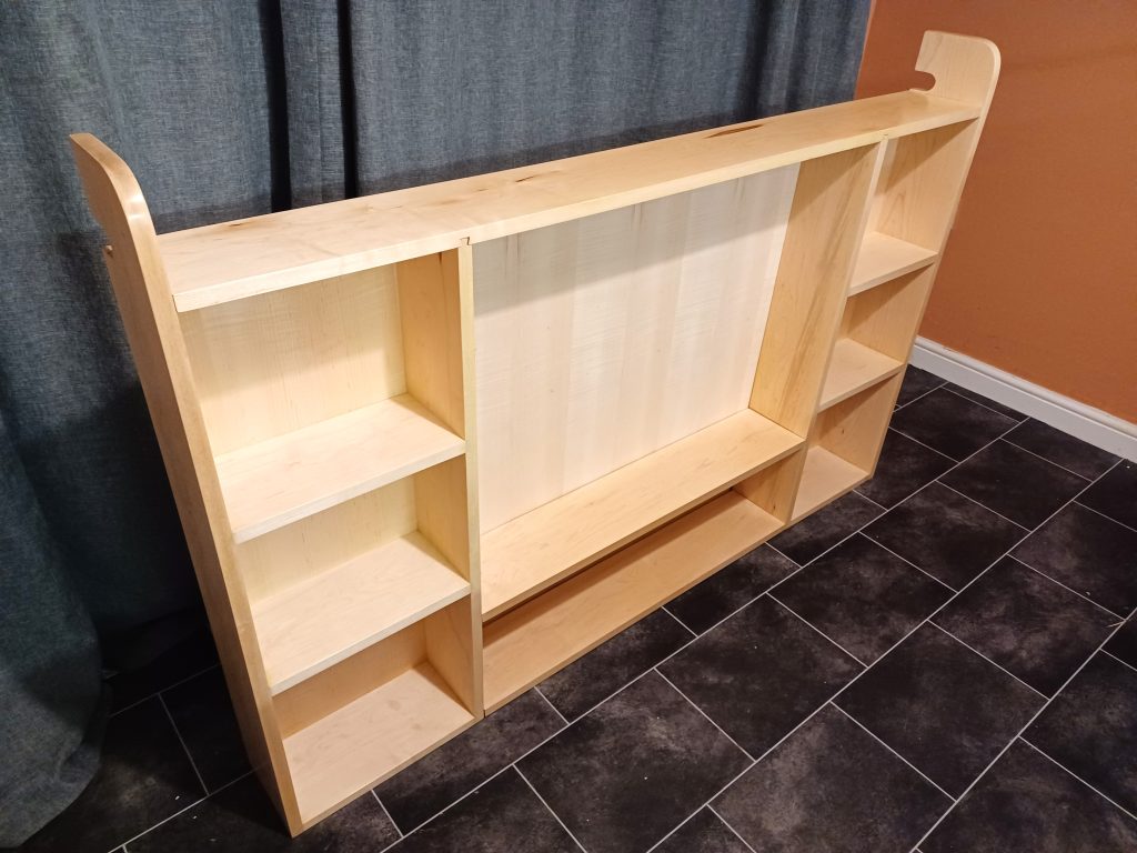 High-angle view of a low, wide wall entertainment unit crafted from light blonde maple wood. The unit features a wide open central compartment backed with a smooth panel, a wide lower shelf, and two vertical side columns with multiple stacked storage cubbies. It rests on a dark tiled floor against a dark blue curtain.