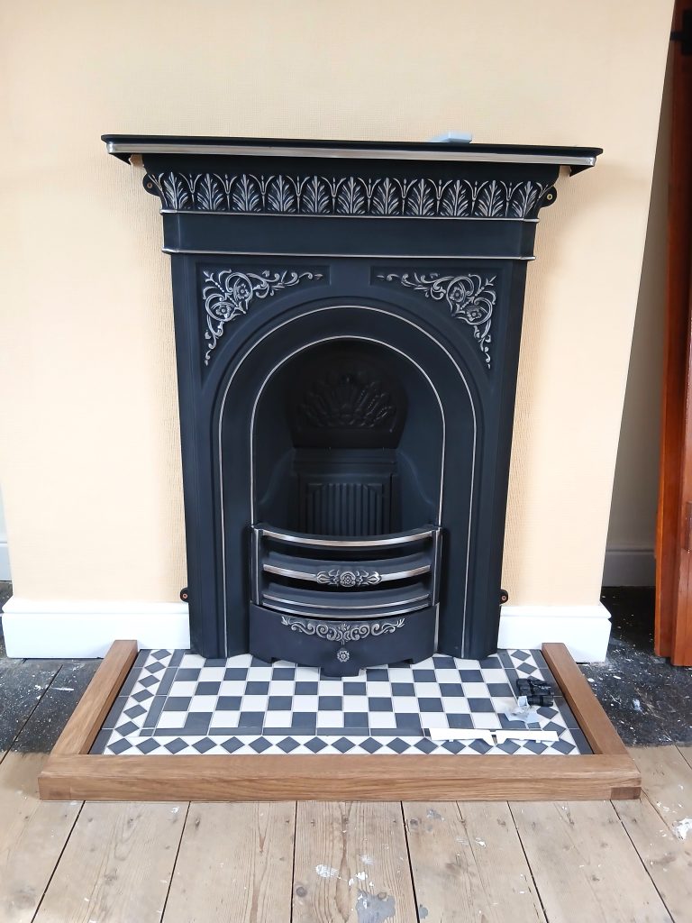 Straight-on view of a low, three-sided solid wooden hearth fender framing a black-and-white checkered tiled hearth floor. Positioned behind it is a decorative, ornate black cast iron fireplace with silver highlighted scrollwork and an arched opening, set against a pale yellow wall.