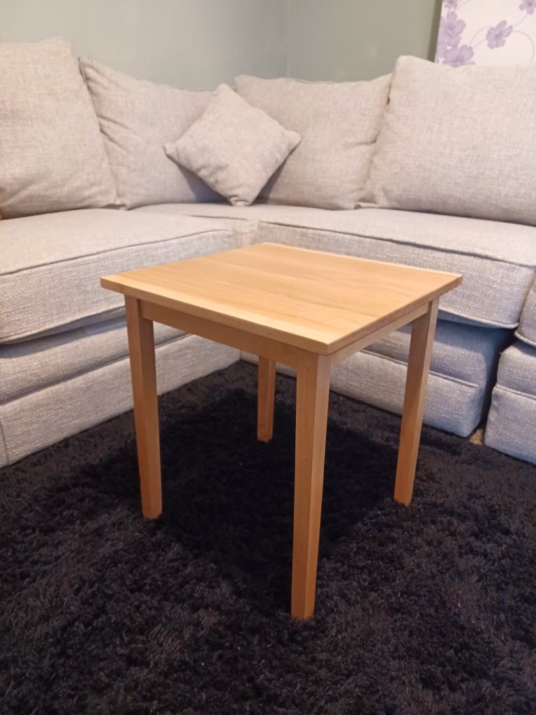 Three-quarter angle view of a small square side table crafted from light maple wood with clean lines and tapered legs. The table stands on a dark shag rug in front of a light grey L-shaped fabric sofa.
