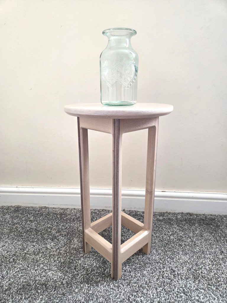 Eye-level view of a tall, slim wooden side table with a circular top made from light maple wood. The four legs feature decorative dark walnut laminate strips running down the outer edges. A clear, embossed glass decorative vase sits centered on the table top against a plain off-white wall background.