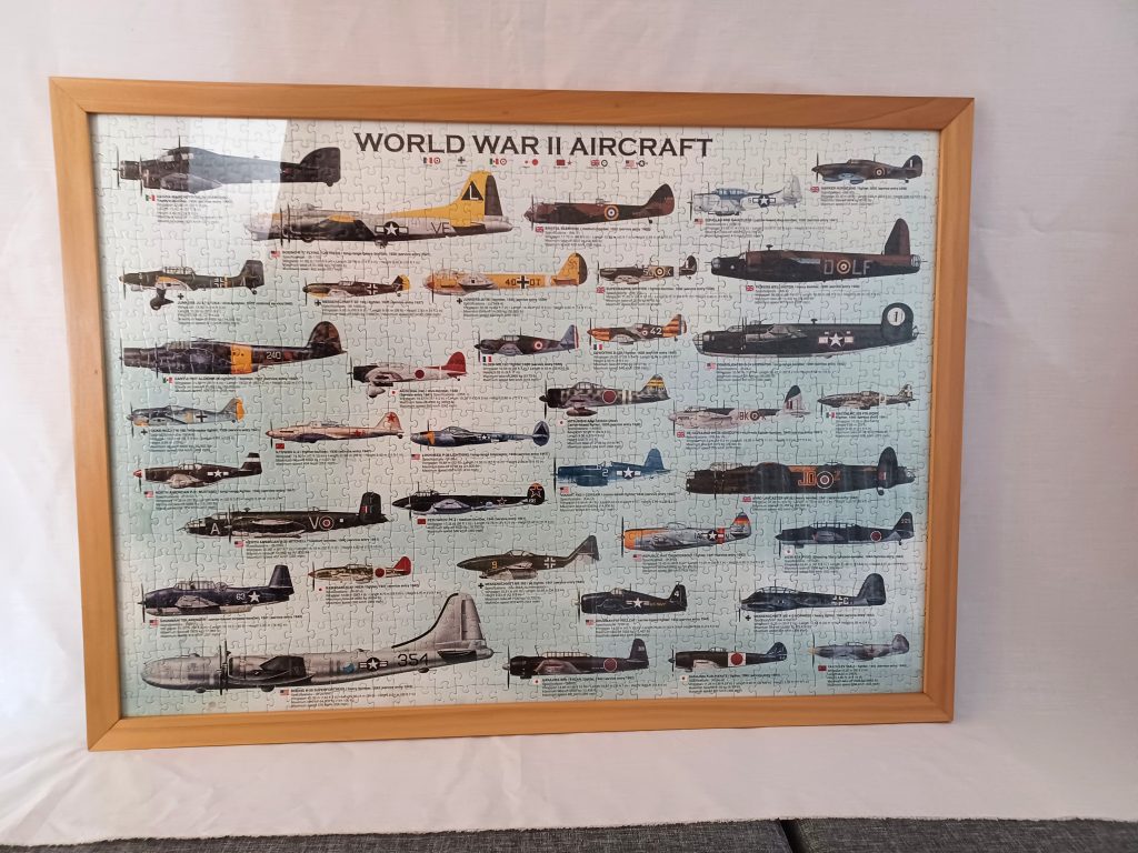 Front-facing view of a wide rectangular light poplar wood frame set against a white backdrop. The frame holds a completed jigsaw puzzle titled "WORLD WAR II AIRCRAFT", featuring detailed illustrations and technical specs of various vintage military airplanes.