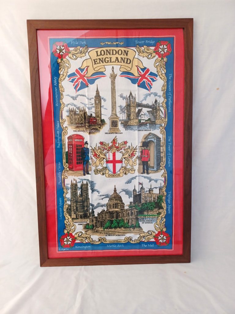 Front-facing vertical view of a rich dark walnut wooden frame set against a white fabric backdrop. The frame encloses a vibrant, vintage-style London souvenirs tea towel with a red border, displaying illustrations of iconic landmarks such as Big Ben, Tower Bridge, St. Paul's Cathedral, a red telephone box, and the London coat of arms.