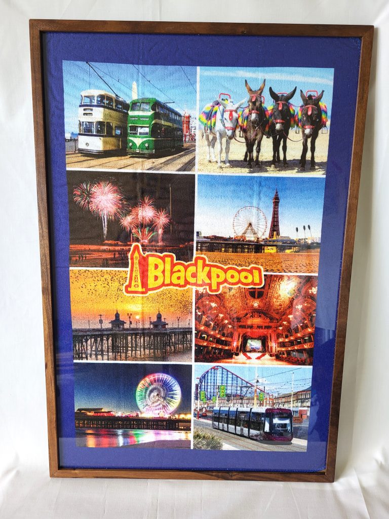 Front-facing vertical view of a handcrafted solid dark walnut wooden frame set against a white fabric backdrop. The frame encloses a vibrant souvenir tea towel with a royal blue border, featuring a collage of Blackpool attractions including historic trams, donkeys on the beach, fireworks, Blackpool Tower, the pier at sunset, and the Pleasure Beach. A red and yellow "Blackpool" title logo is prominent in the center.