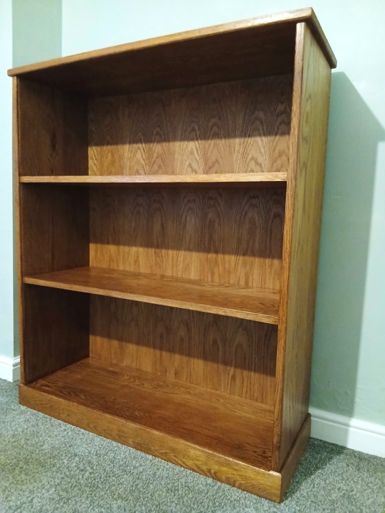 Three-quarter angle view of a medium-height, three-shelf solid oak bookcase finished in a warm medium-oak stain. The unit features visible natural wood grain patterns across the back panel and shelves, standing on a grey carpeted floor against a light green wall.