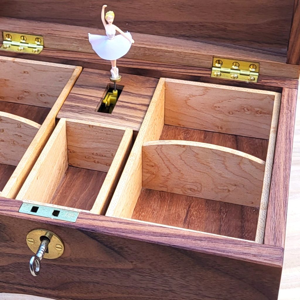Close-up view of an open handcrafted wooden music box featuring rich dark wood outer walls and light-toned figured wood interior compartments. Inside, a miniature ballerina in a white tutu stands poised on a central music box mechanism. The box includes solid brass hinges, a brass escutcheon plate, and a key inserted into the front keyhole.