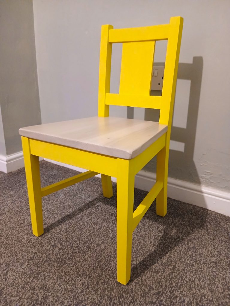 Three-quarter angle view of a small wooden child's dining chair resting on a grey carpeted floor against a light grey wall. The chair features a sturdy frame and vertical backrest painted in bright yellow, paired with a smooth, natural light wood seat top.